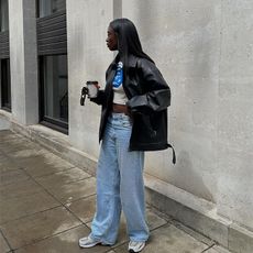 woman wears crop top and baggy light wash denim jeans and sneakers with an oversized leather jacket while holding a to go coffee cup