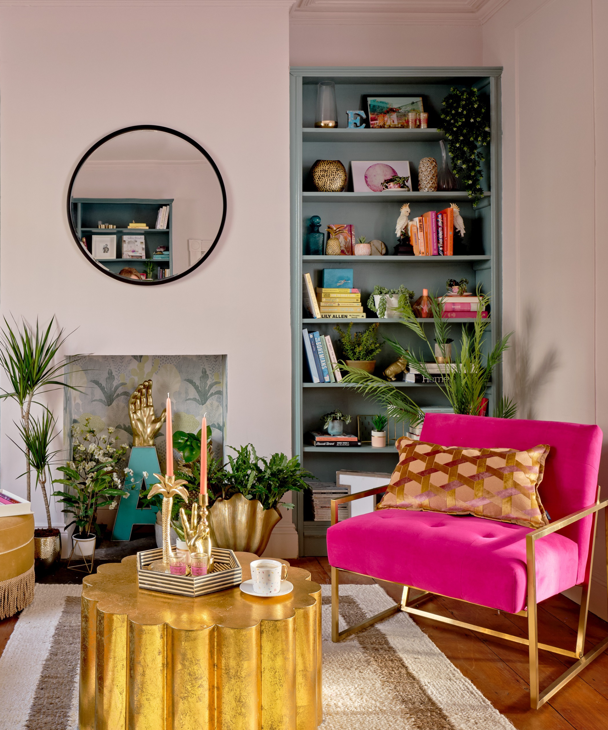 Corner of a living room with a blue book case in fireplace alcove, a pink chair, a gold coffee table, and a wallpapered fireplace