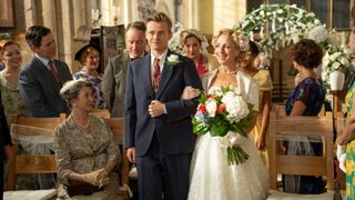 Eddie Devine (Barney Wilkinson), wearing a navy blue suit, walks his mother Mrs Devine (Claudie Blakley) up the aisle on her wedding day. The seats are full of guests smiling at them, and there is a floral arch behind them. Mrs Devine is in an ivory wedding dress and holding a bouquet of white flowers with dashes of red and blue.