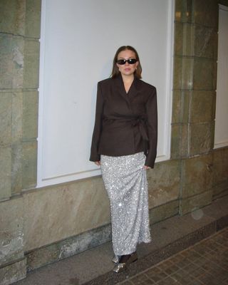 Dina wears sunglasses, a brown wrap blazer, silver sequin skirt, and metallic silver flats.