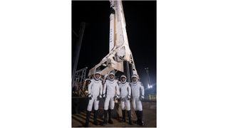 four astronauts and white and black spacesuits stand in front of a rocket on the launch pad at night.
