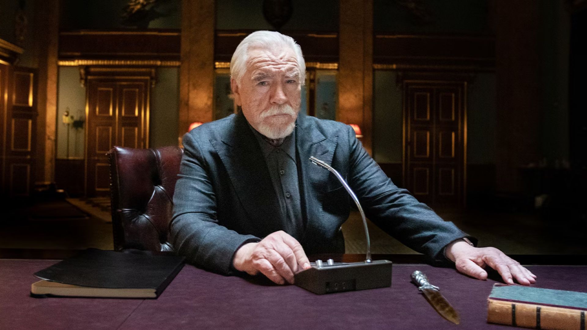 Brian Cox sits at a desk pressing a button on an intercom