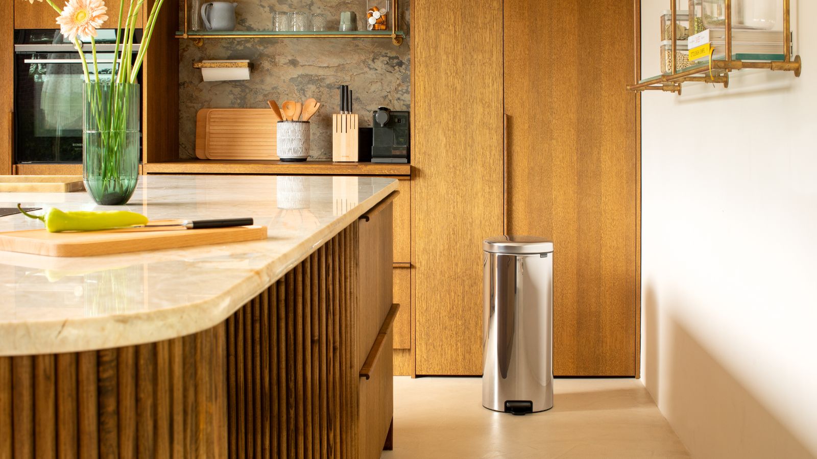 A wooden kitchen with wooden cabinets, a wooden panelled island with marble countertop, gold shelving and brown marble backsplash. A silver pedal bin is seen in the background.