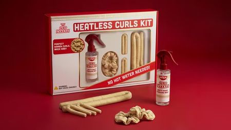 A Cup Noodles branded "Heatless Curls Kit" featuring a box and loose hair accessories like a spray bottle labeled "Broth Boost Finishing Mist," scrunchies, and foam rollers designed to look like ramen noodles against a solid red background.