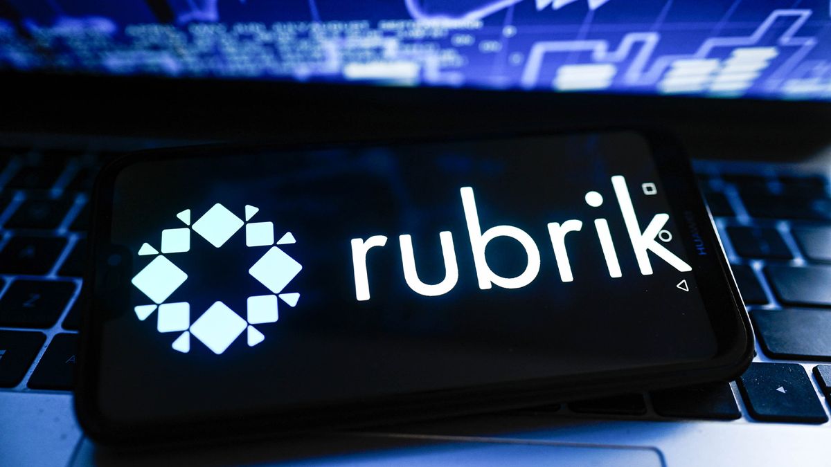 Rubrik logo and branding displayed on a smartphone sitting on a laptop with screen showing stock market fluctuations.