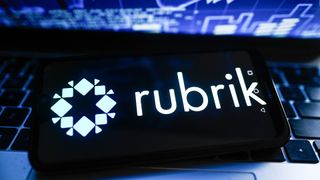 Rubrik logo and branding displayed on a smartphone sitting on a laptop with screen showing stock market fluctuations.