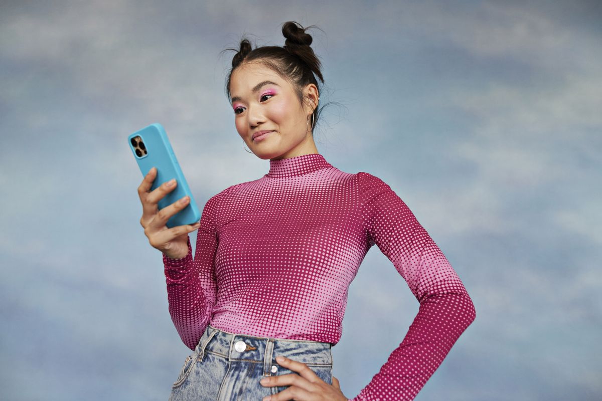 Feminine-presenting Gen Z person wearing a dotted pink top, with pink eyeshadow and space buns, holding a blue smartphone