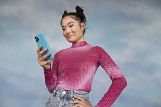 Feminine-presenting Gen Z person wearing a dotted pink top, with pink eyeshadow and space buns, holding a blue smartphone