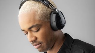 A man with bleached blond hair wearing a pair of Skullcandy Aviator 900 ANC headphones in front of a plain gray background