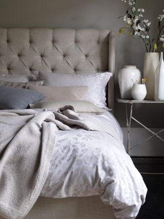 neutral bedroom scheme with button backed upholstered bed, vases on vintage side table, damask style bedding, textured throws and pillows