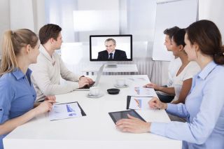 Five people with laptop and tablet computers watch a man speaking on a video display.
