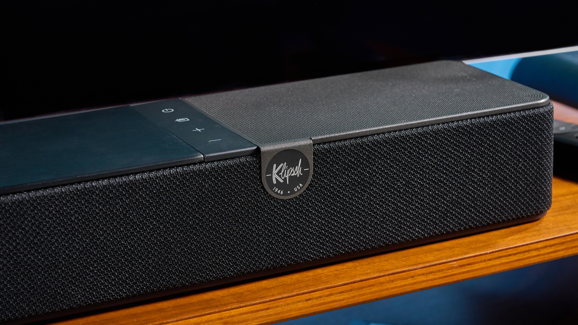 the klipsch flexus core 100 soundbar photographed on a wooden tv stand with a tv behind, showing the remote, buttons, and inputs