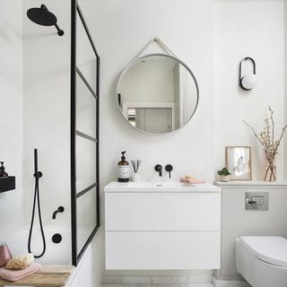 White bathroom with white vanity below round mirror next to bath and toilet