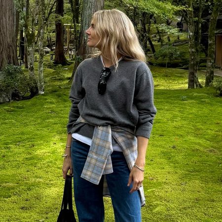 British Lucy Williams wearing a casual-cool outfit idea while visiting Japan wearing a gray sweater layered over a white long-sleeve T-shirt, a plaid shirt wrapped around her waist, and jeans