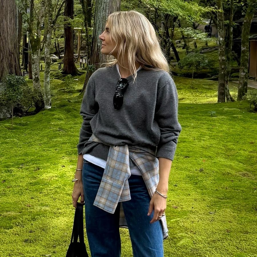 British Lucy Williams wearing a casual-cool outfit idea while visiting Japan wearing a gray sweater layered over a white long-sleeve T-shirt, a plaid shirt wrapped around her waist, and jeans