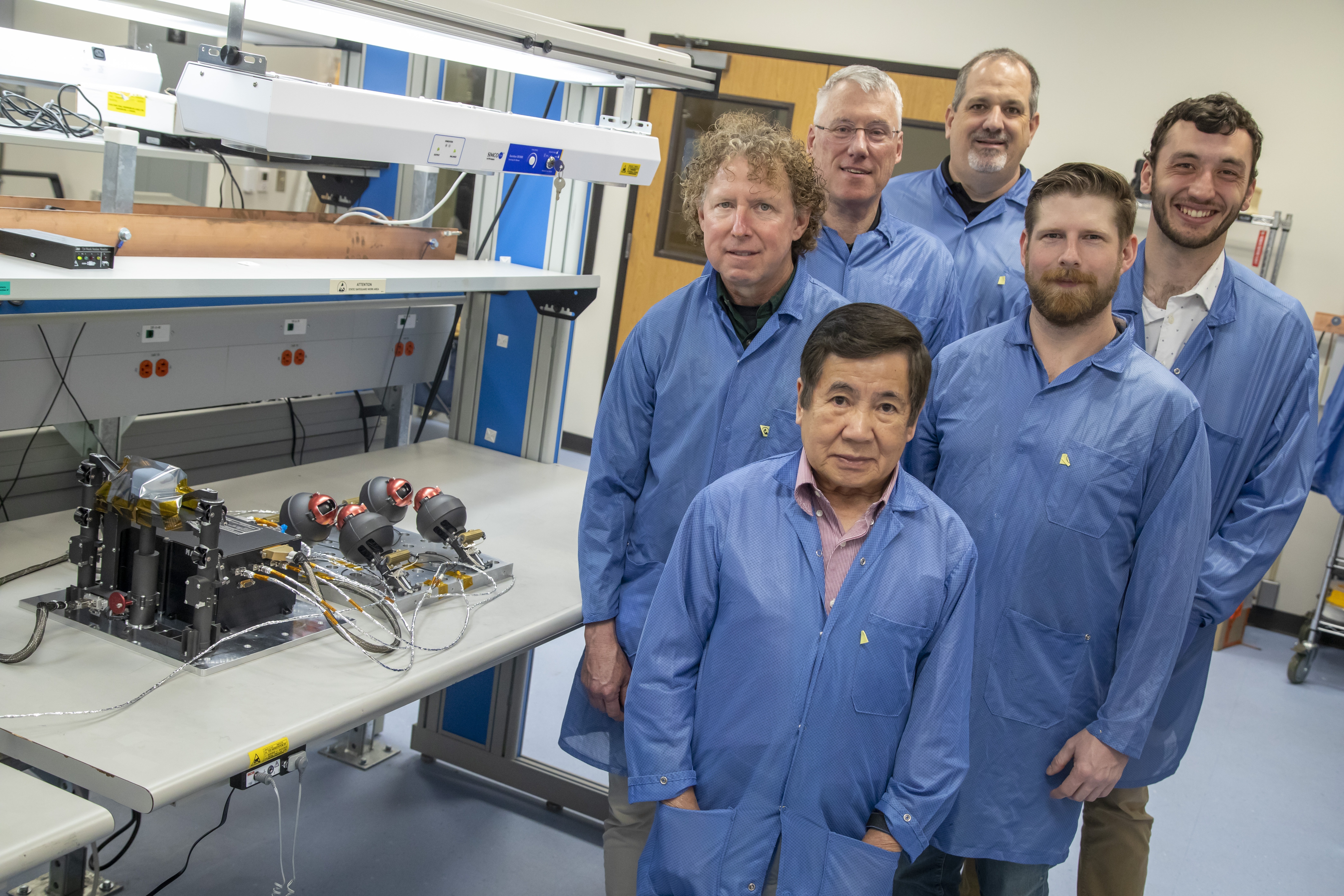 A group of people wearing blue coats stand next to a laboratory bench.