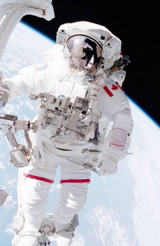 chris hadfield in spacesuit floating in space with the earth behind. a canadian flag is on his spacesuit shoulder