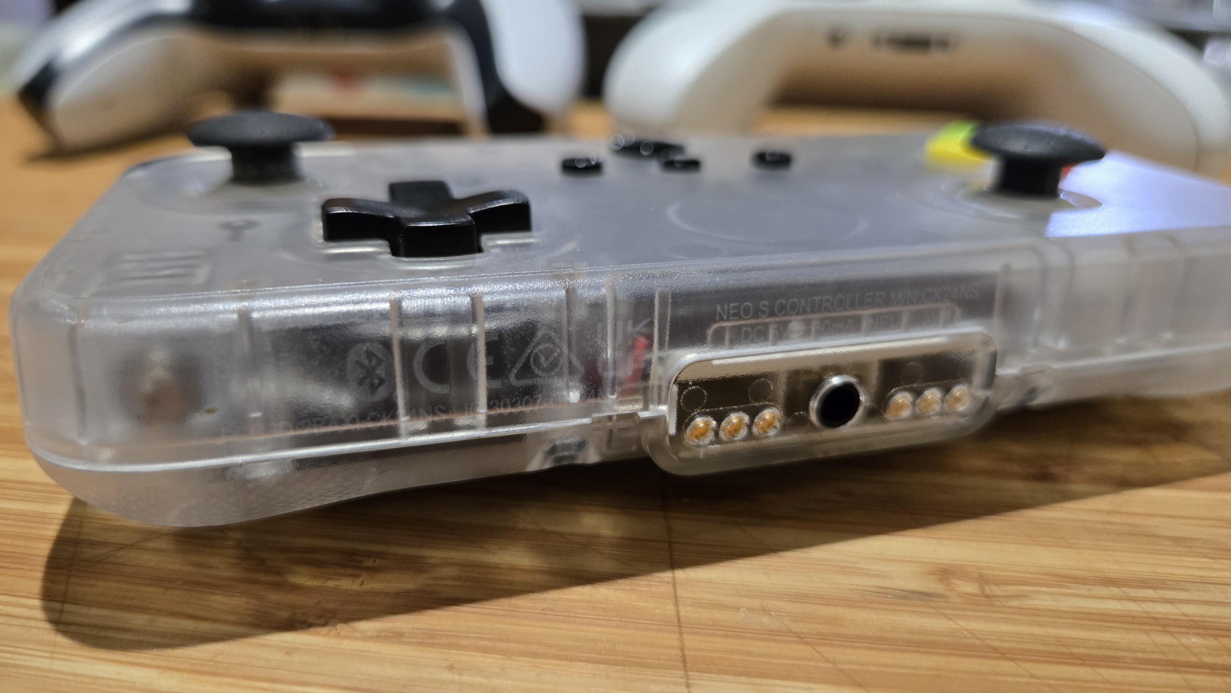 A Classic Clear edition of the CRKD NEO S rectangular controller sits on a desk, with a DualSense PS5 controller and a Xbox controller vaguely visible behind it. The perspective of the image is quite dramatic, with the CRKD controller dominating the frame, but one still gets a sense of how much smaller it is compared to the other two controllers. The body of the controller is made of a cloudy clear plastic that allows the viewer to see the internals.