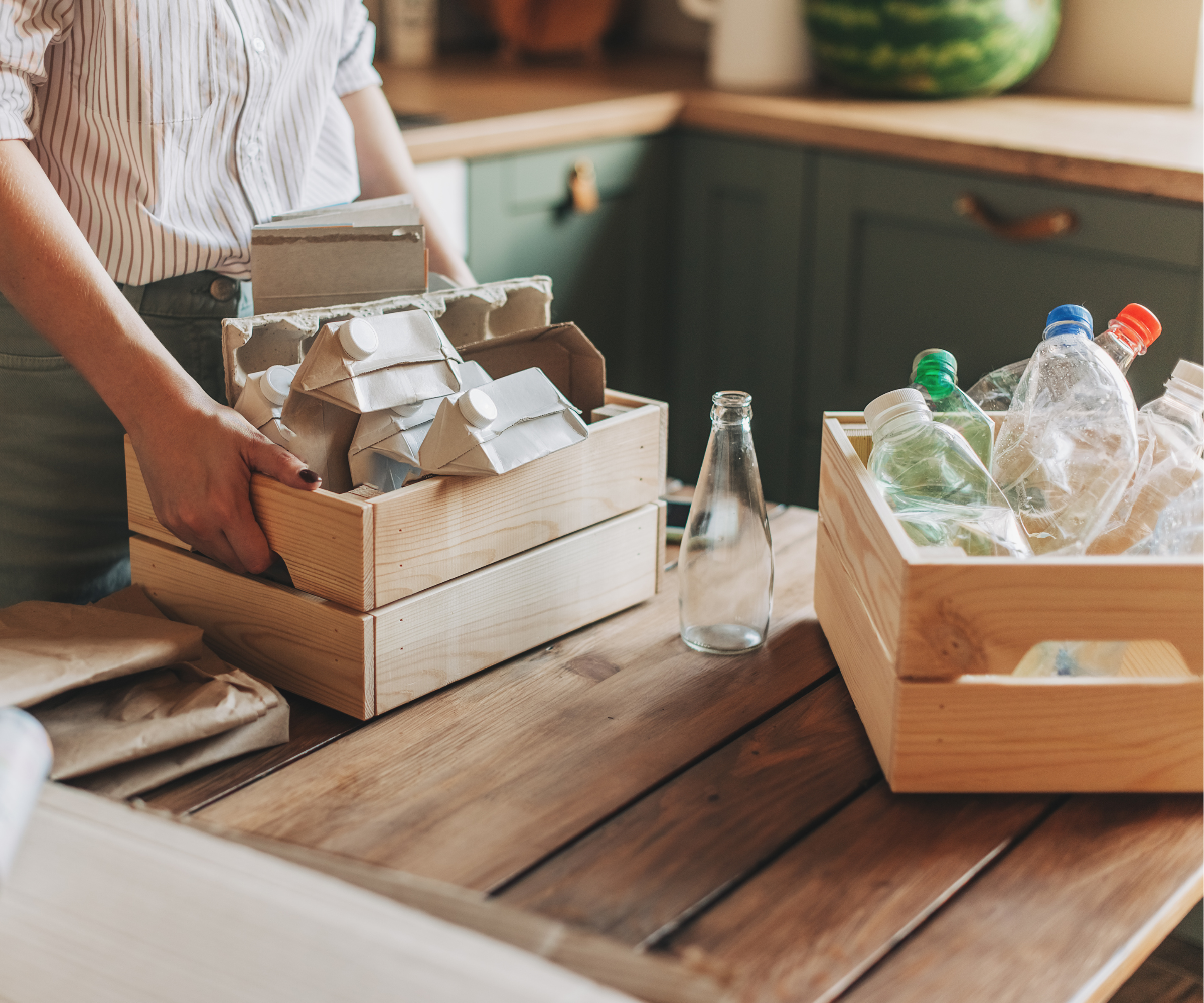 Person holding a crate of cardboard recycling on a wooden table which has another crate of plastic recycling and a single jar in the middle