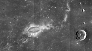 A black and white image of the moon&#039;s surface. A large white pattern is seen in the middle-left and there are craters toward the right.