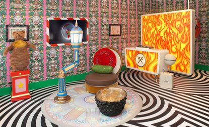Always Close installation by Studio Job in Luxembourg. A very colourful room with a burger shaped chair, a rock shaped coffee table, a bar counter with flames printed on it, a teddy bear on a side table and a swirling black and white floor.