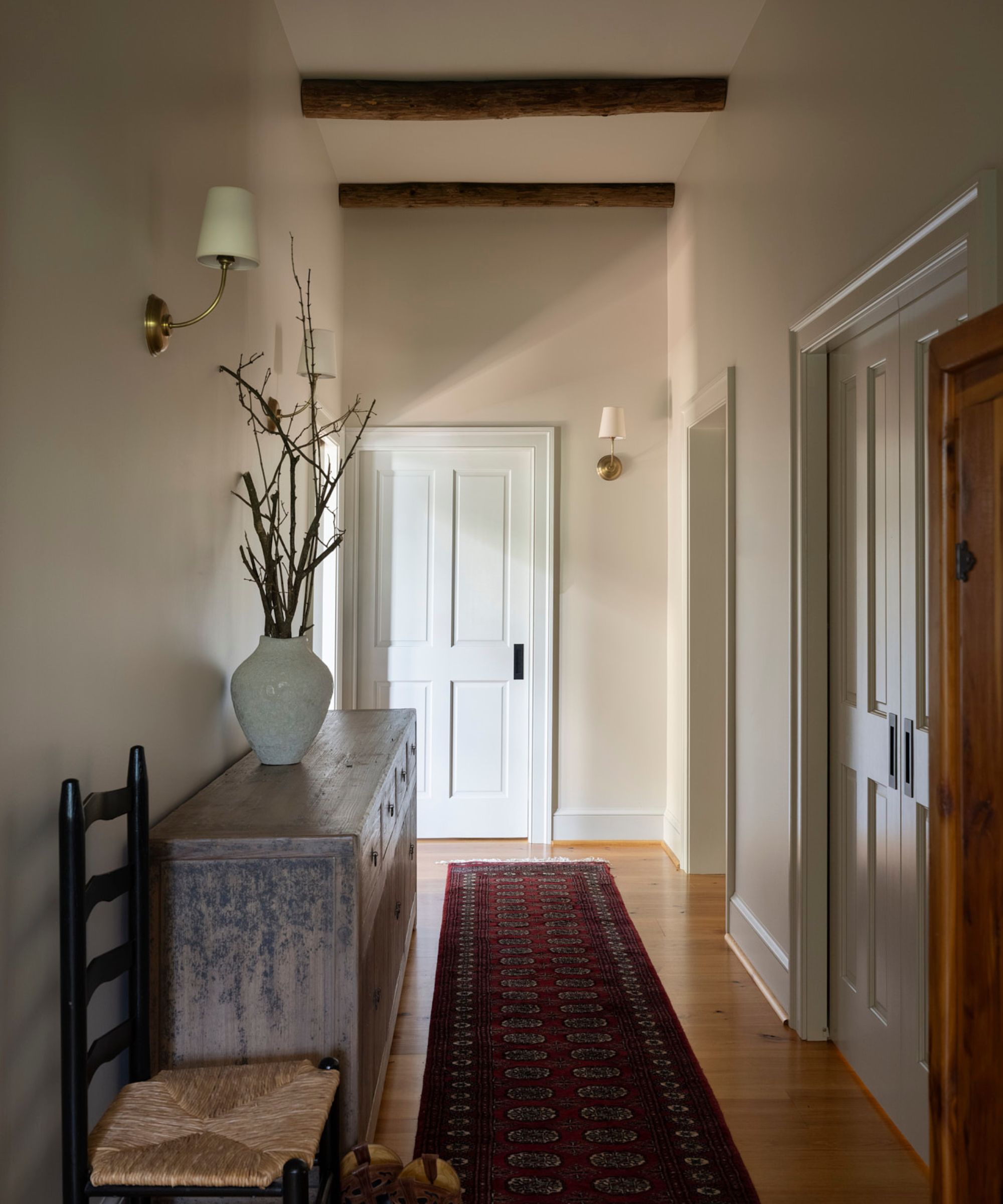 A hallway with off-white walls, wood ceiling beams, wood flooring with a dark red runner rug, a vintage wicker chair, and a wooden console table with a vase of branches