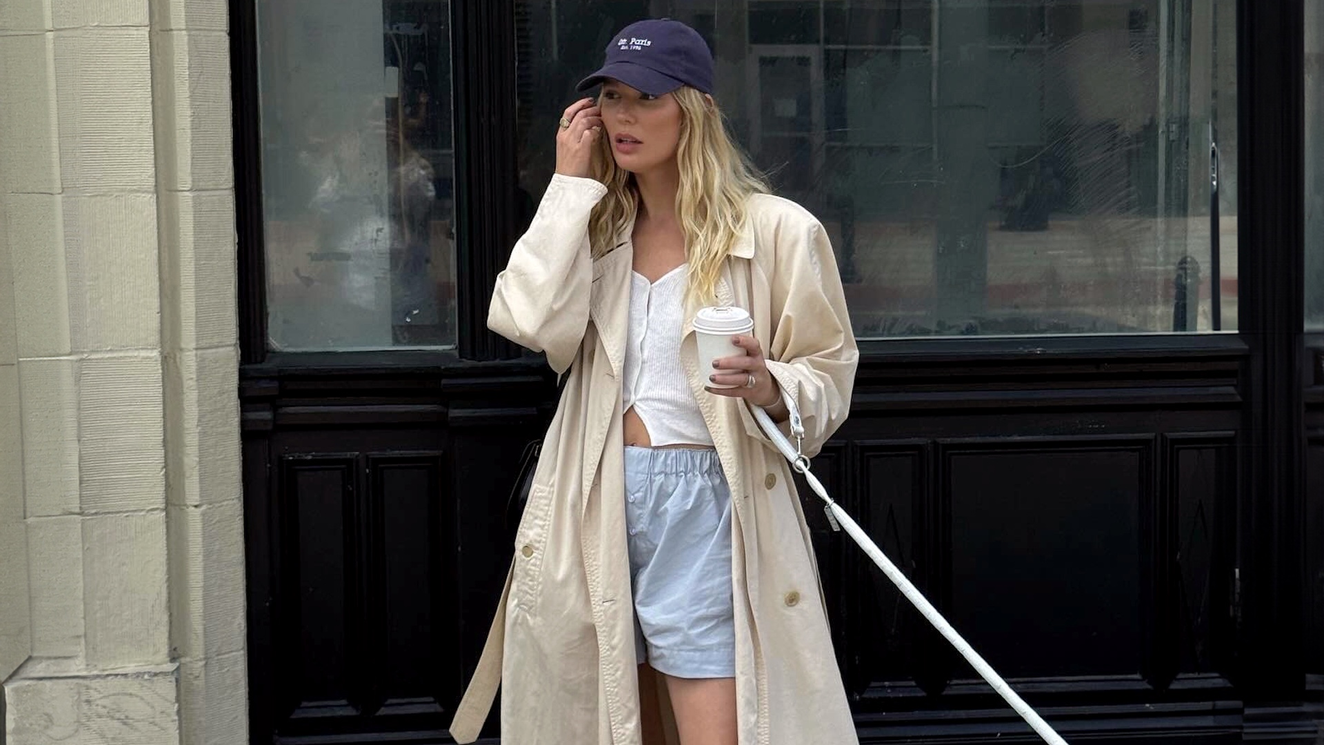 style influencer Megan Adelaide Vega poses while walking her dog in a dark blue baseball cap, long trench coat, white button-down knit top, and blue boxer shorts
