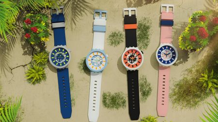 Four Swatch watches in pink, blue, black and white against a background with leaves on