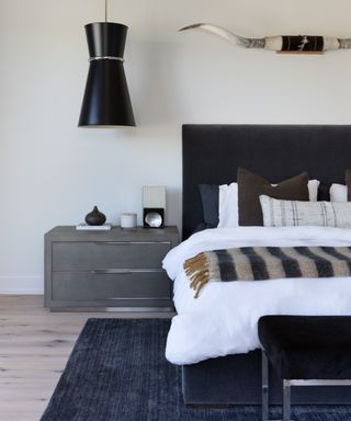 blue and white modern bedroom with velvet bed, grey nightstand, blue rug, black statement pendant, cow horns above bed, stripe blanket, brown, blue and white pillows, metal and velvet stool