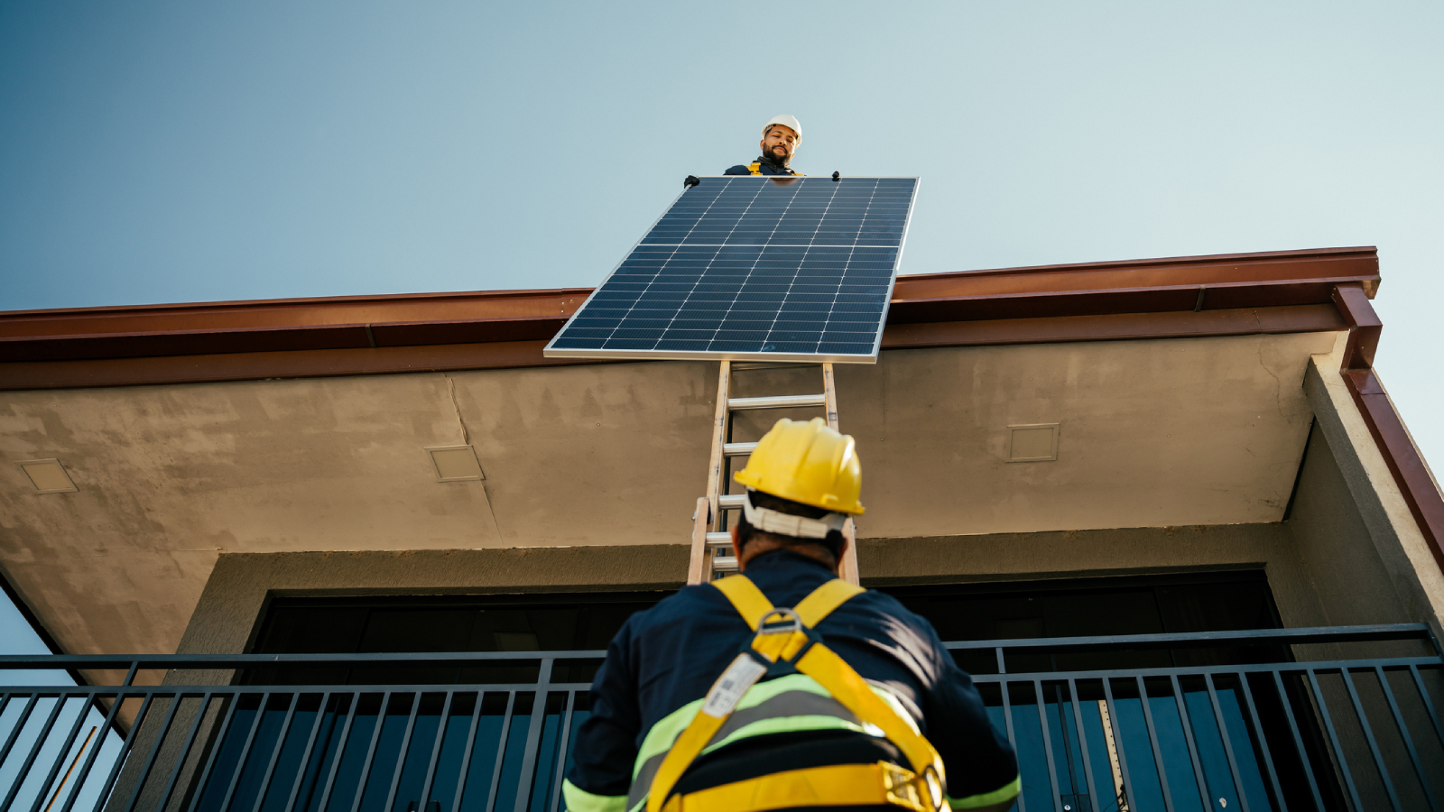 solar panel installer handing solar panel from roof to man on ladder both wearing PPE