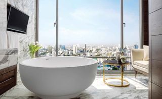 White bath, coffee table and chair in front of full-height windows overlooking Bangkok