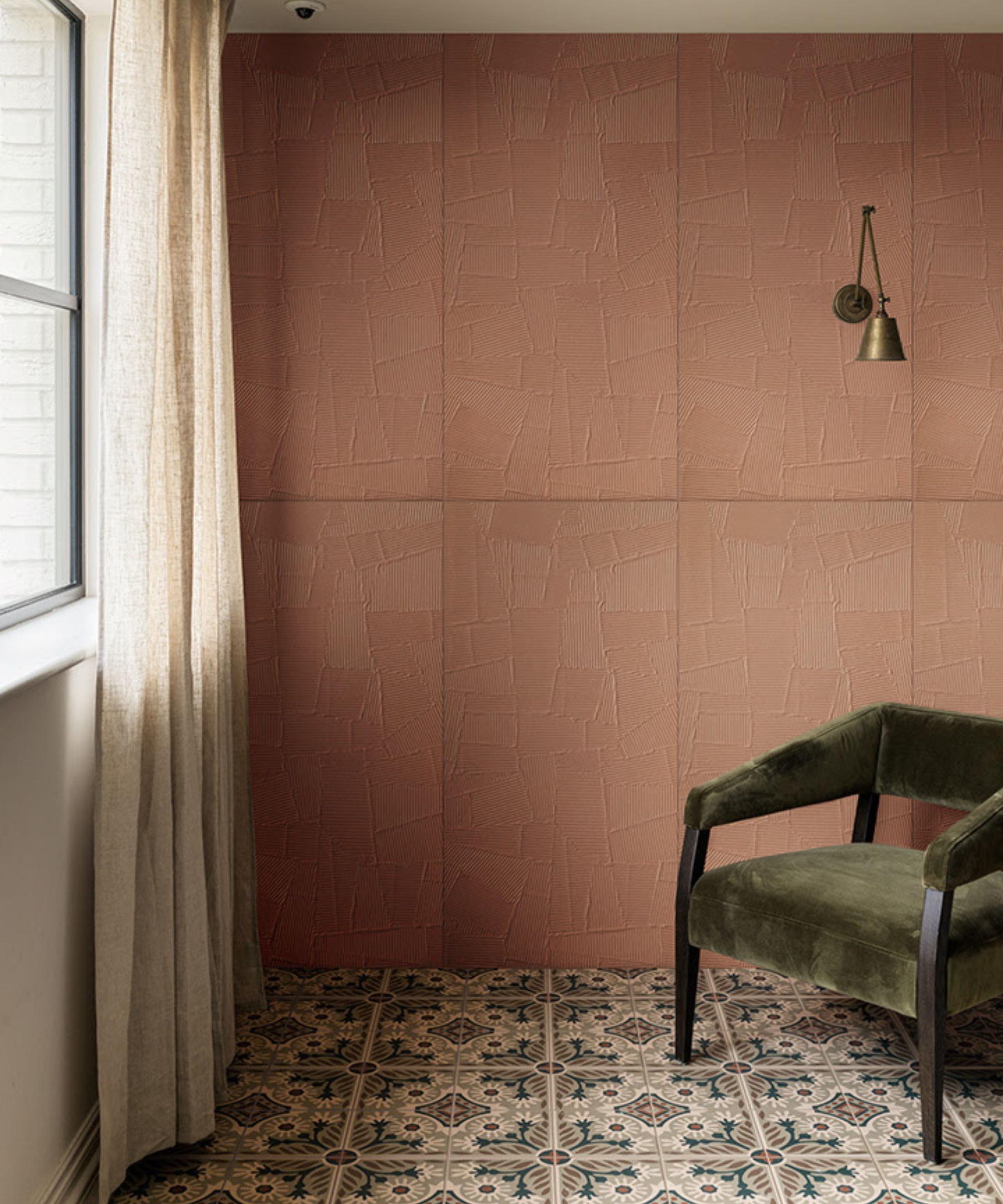 Warm terracotta feature wall with textured plaster finish, paired with patterned ceramic floor tiles in muted blue and cream. A single green velvet armchair sits to the right, beside a brass wall light, with soft daylight filtering through a curtained window.