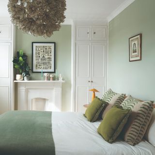 A light sage green-painted bedroom with a large floral ceiling light and green cushions and throw on the bed