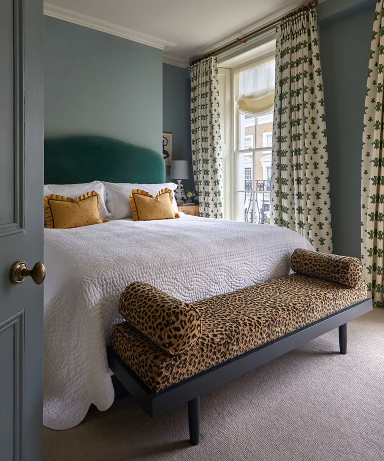 green bedroom with large green velvet headboard and a leopard print upholstered ottoman at the foot of the bed