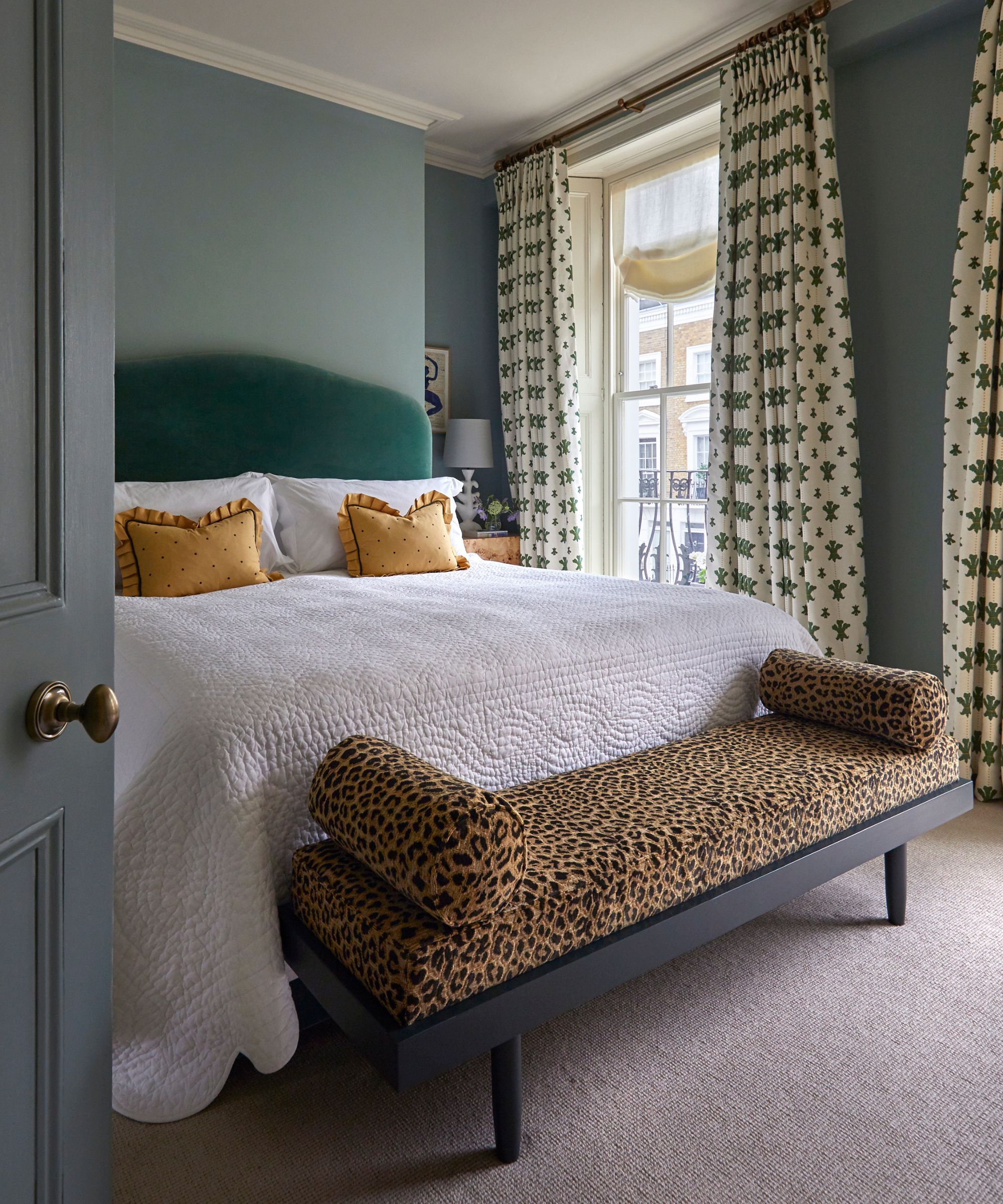 green bedroom with large green velvet headboard and  a leopard print upholstered ottoman at the foot of the bed