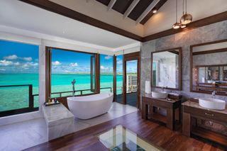 A large standalone bathtub in a bathroom with double sinks and huge glass windows overlooking the ocean.