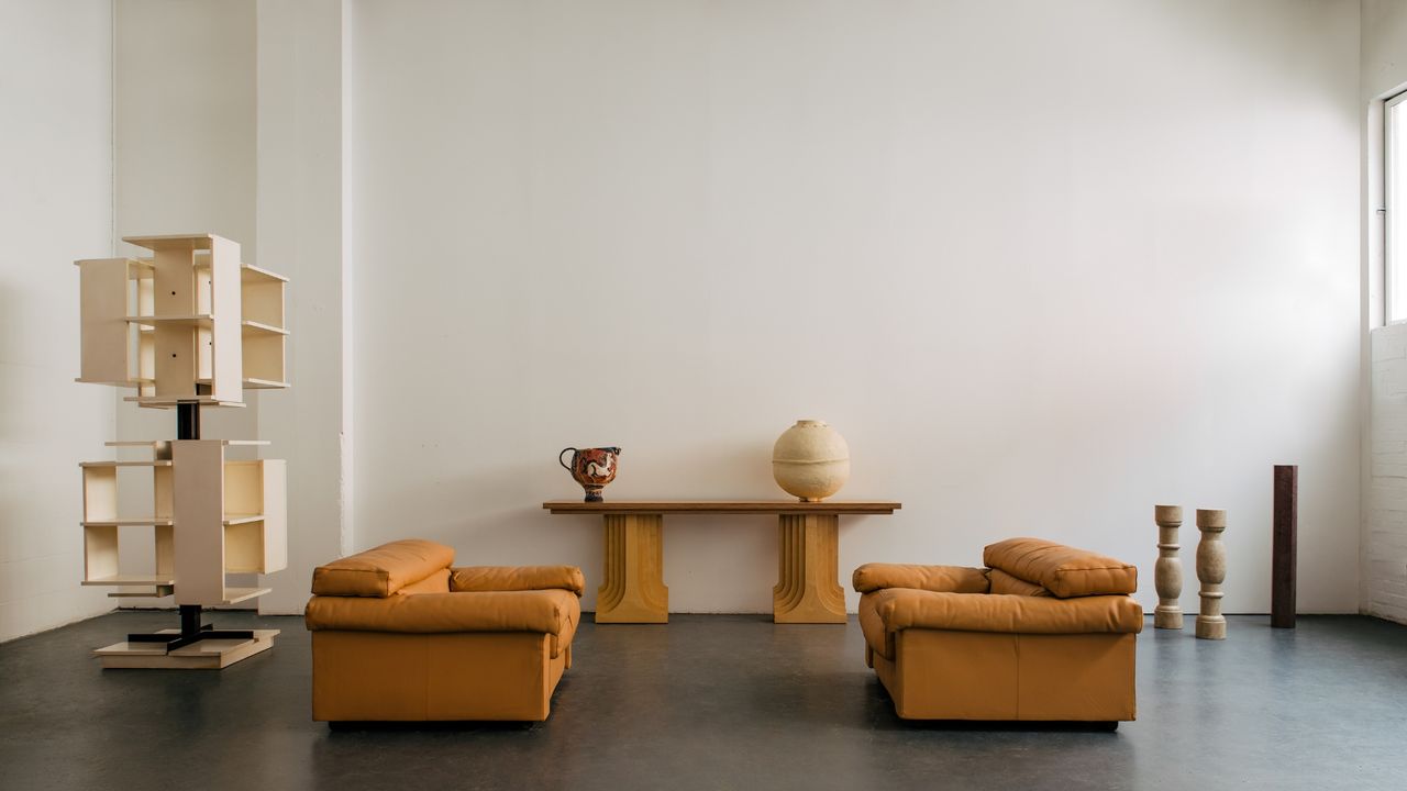 An industrial studio with black floors and white walls features a selection of modern design pieces ranging from mid-century modern orange sofas, positioned in front of each other, to sculptural bookcases, wooden objects, and tables.