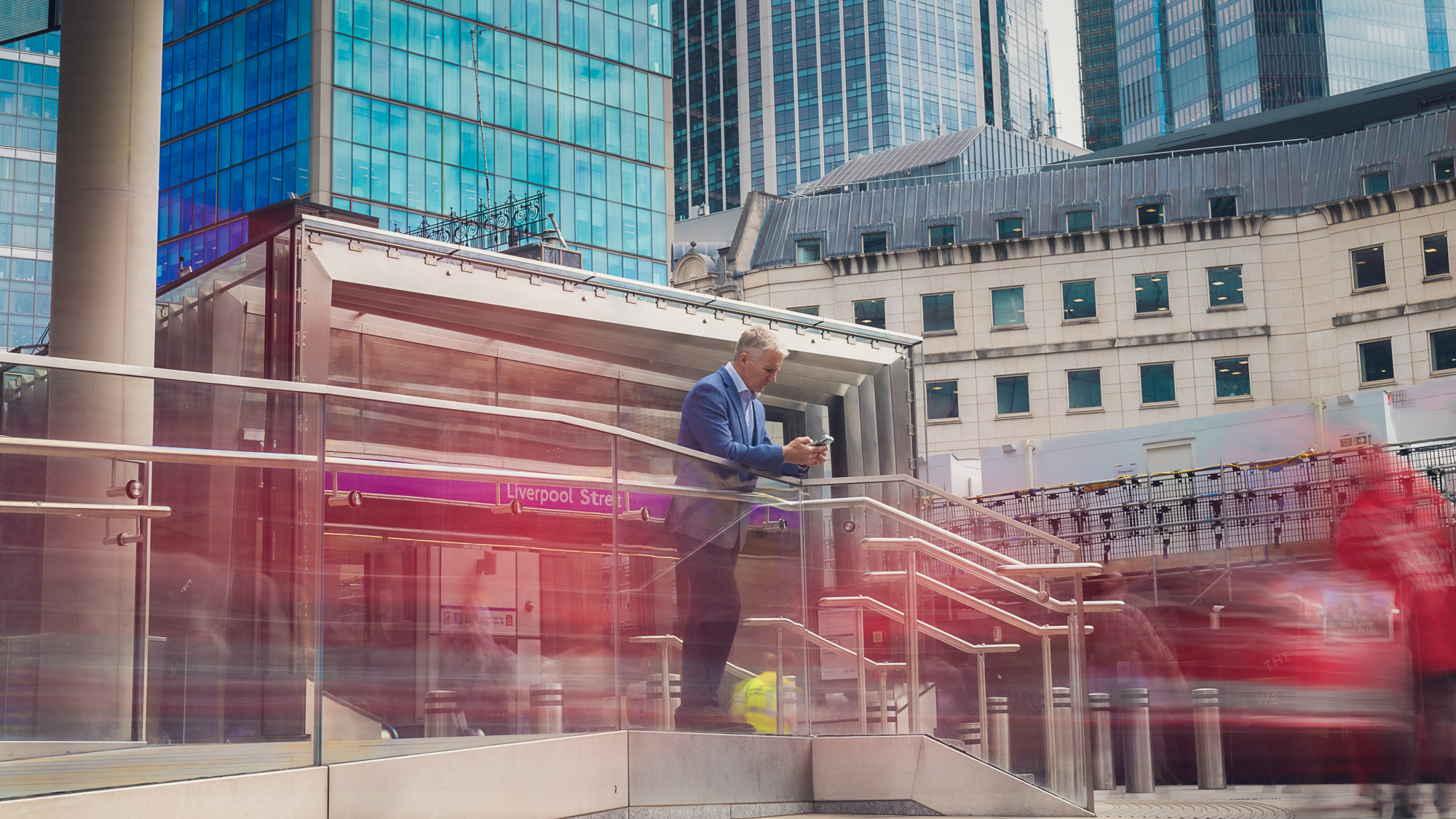 A man stands still beside a railing in an urban setting, while a long shutter speed has been used to blur people around him into a colorful blur