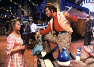 a girl in a red check dress speaks to a man working at a carnival in Carousel