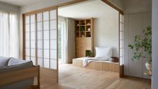 A Japanese living room with a vase of stems on a side table, a gray couch, a sliding rice paper screen, and a bench with a pillow and a display case of ceramics