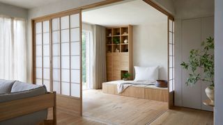 A Japanese living room with a vase of stems on a side table, a gray couch, a sliding rice paper screen, and a bench with a pillow and a display case of ceramics