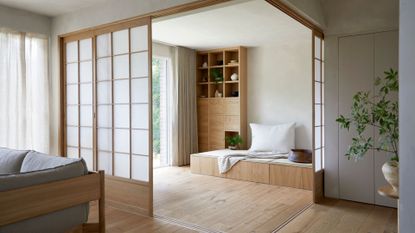 A Japanese living room with a vase of stems on a side table, a gray couch, a sliding rice paper screen, and a bench with a pillow and a display case of ceramics