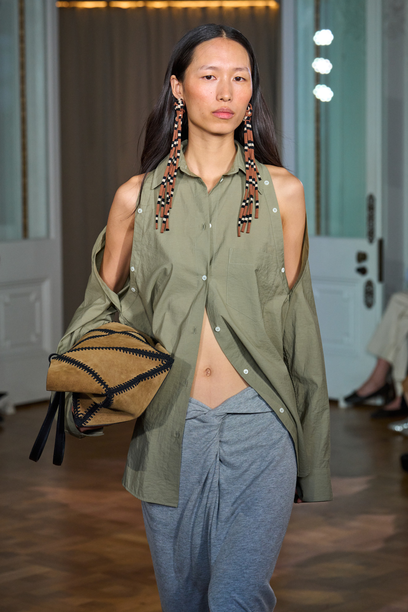 A model at the Nanushka SS26 show with loose waves, minimal makeip and wearing long patterned earrings and a green, cut-out shoulder shirt.