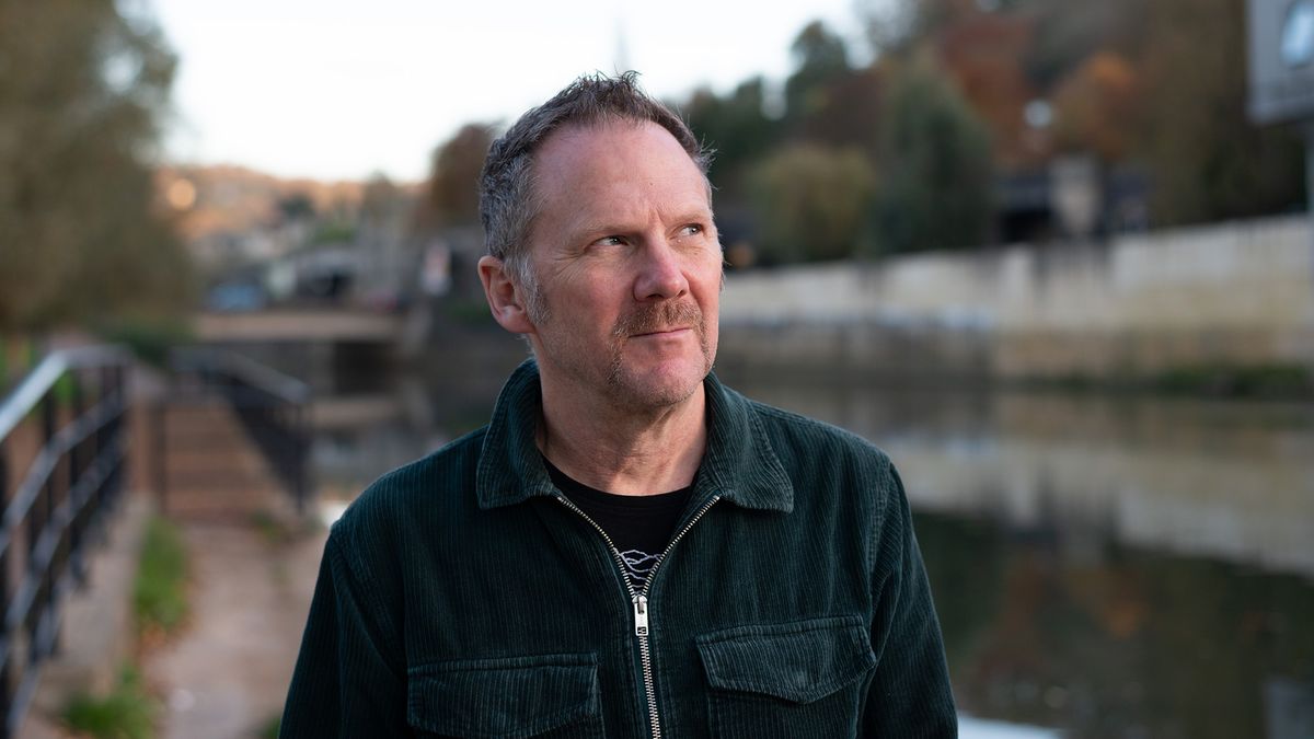 Man with green jacket and handlebar moustache, stood in front of river