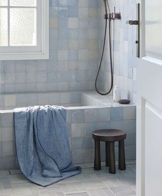 blue tiled bathroom with blue denim colored towel draped over the bath, timber stool beside