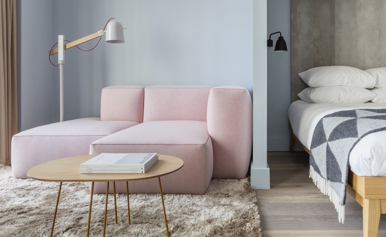 A room in the Leman Locke hotel. Pale blue walls, with a dusty pink couch in the corner and a wooden table. To the right, we see a part of the bed.