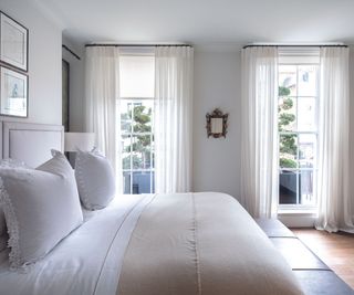Double bed in pale grey bedroom with wooden floor, cream and white bedding and white curtains dressing floor to ceiling windows. A renovated four storey London townhouse, home of Louise Bradley, interior designer.
