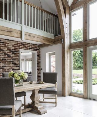 The interior of an oak frame home with a large wall of glazing, a white tiled floor and a wooden table and chairs with hydrangeas in a vase on it