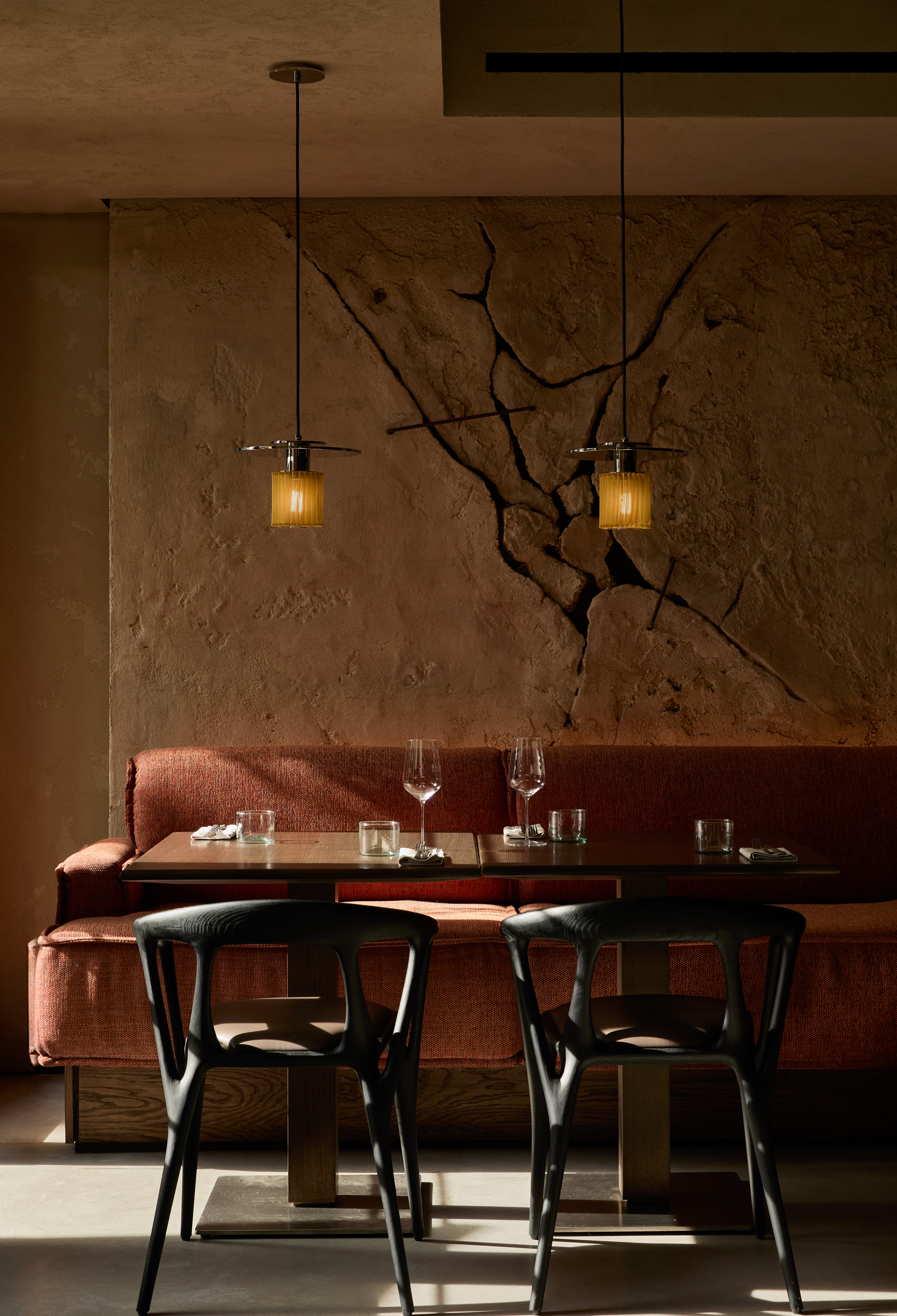 A restaurant dining room brought to life by industrial-style, metal and fabric pendant lanterns, dark wood furniture, and upholstered clay-orange banquette features cracked cement walls with a striking effect.