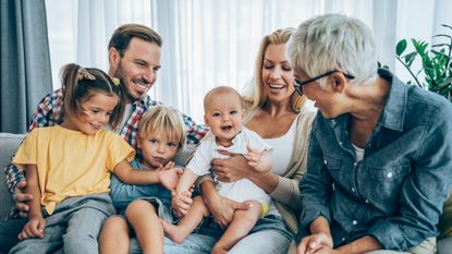 A grandmother smiles at her three young grandchildren. Her daughter and son-in-law hold the children. They are all sitting on a couch, looking happy.